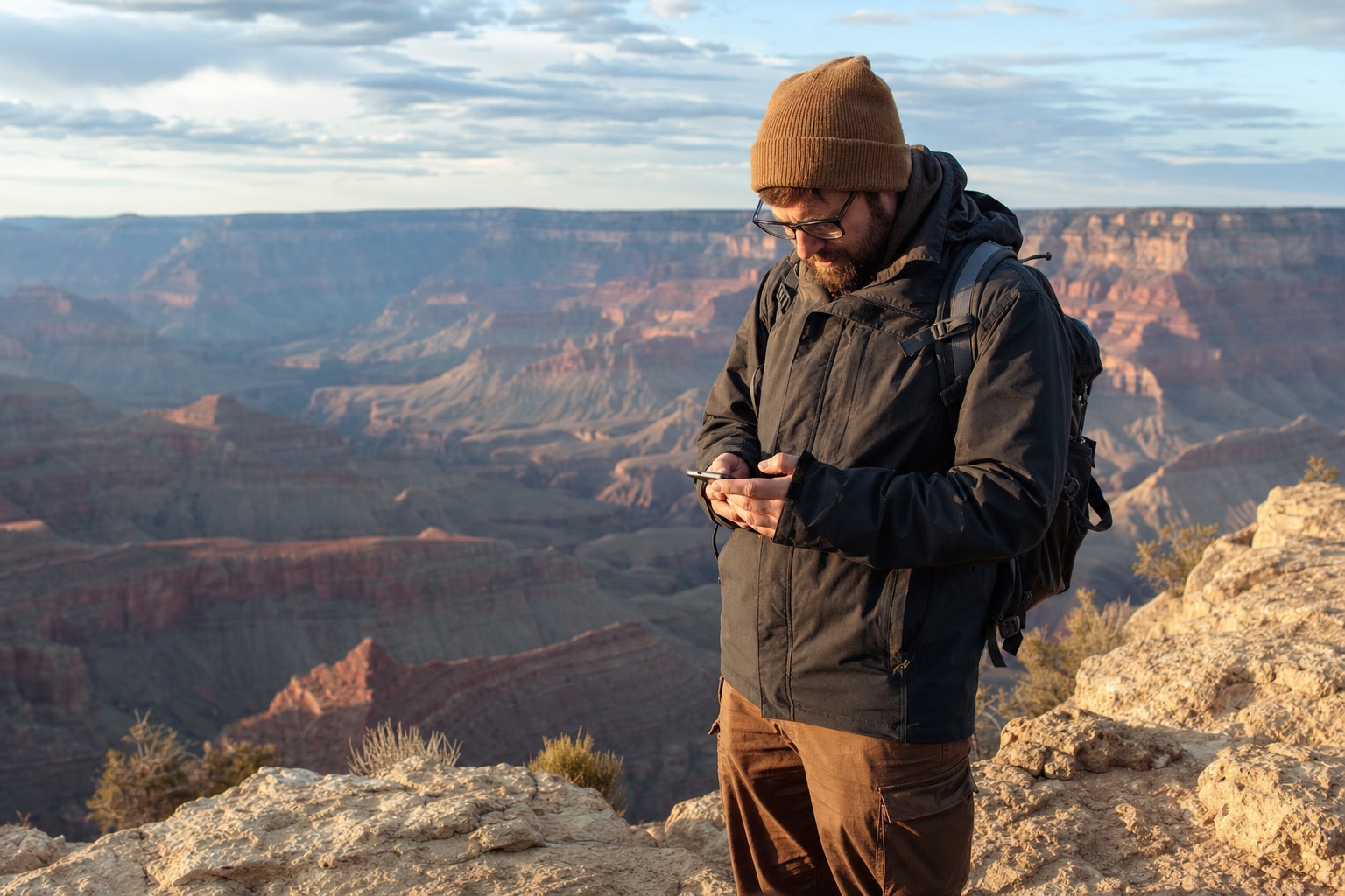 A traveler checking a phone at a Grand Canyon rim pullout with the gorge stretching behind in afternoon light