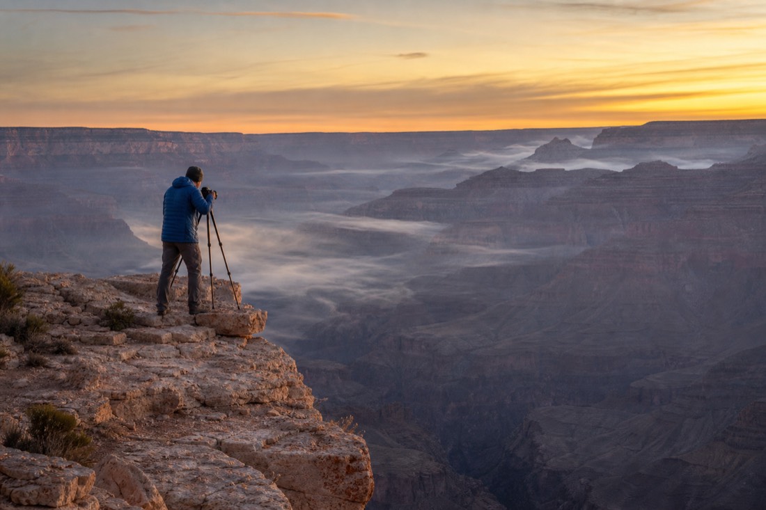 A lone photographer with a tripod at the canyon edge at dawn with mist hanging in the gorge below