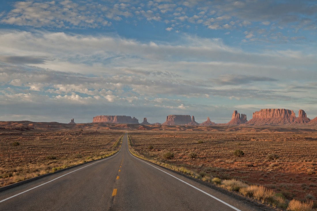 A highway stretching across empty desert toward distant mesas under a huge afternoon sky
