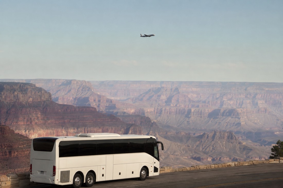 A white tour coach parked at a canyon overlook while a small airplane passes above the rim in the distance